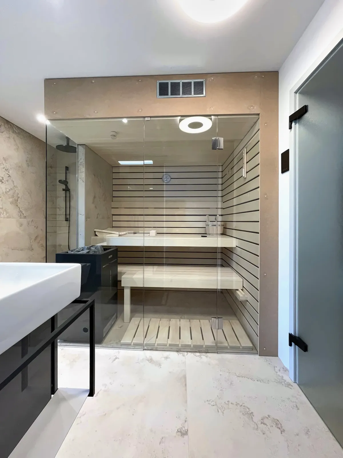 Sauna hinter Glas, bodenebene Dusche und Waschplatz in einem Raum. Grossformatige Keramik in Steinoptik, eine Wand in Petrol.