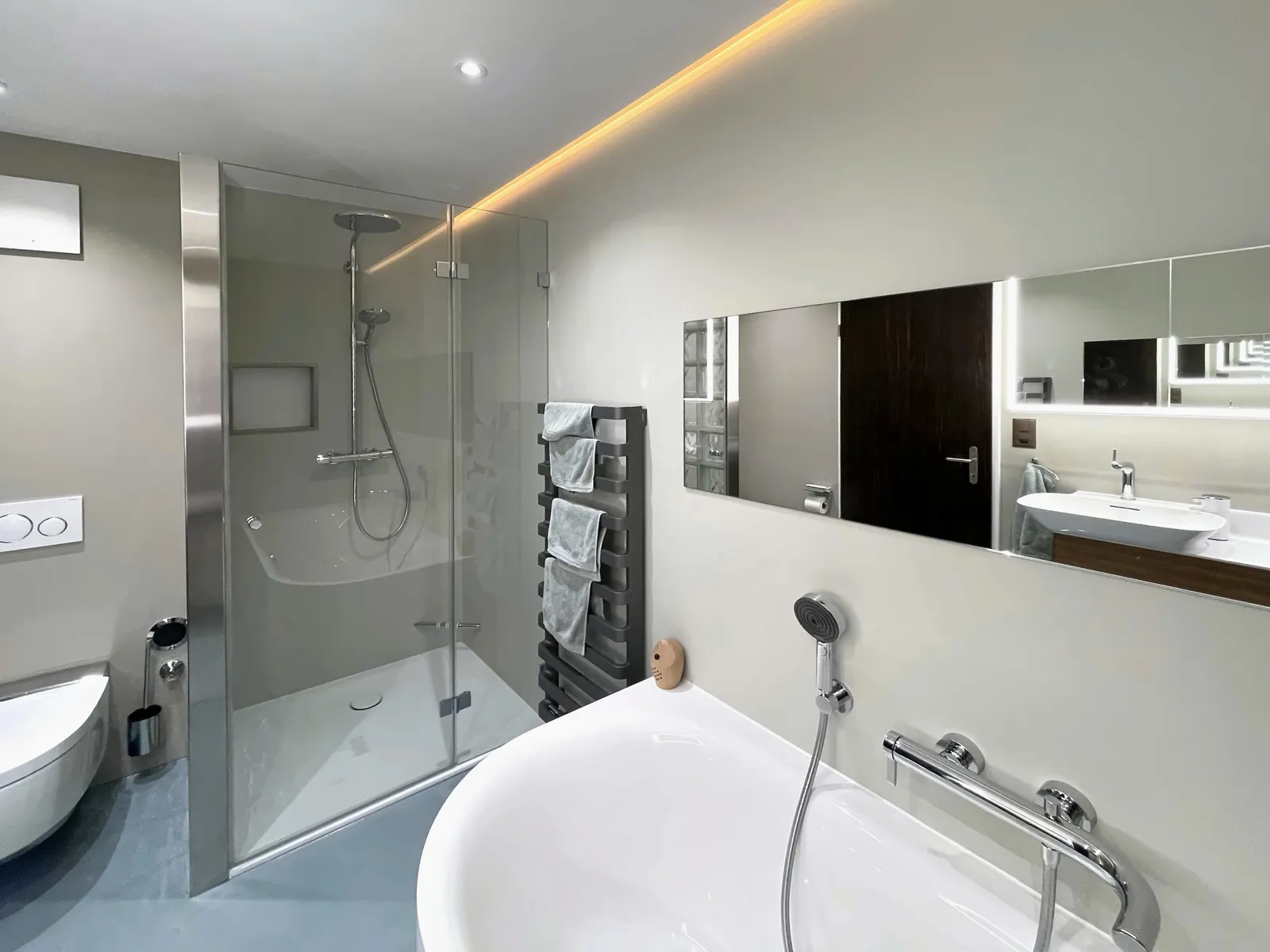 Wanne, Dusche hinter Glas und ein Waschtisch in Holz. Eine Lichtvoute zeichnet die Decke nach, der Boden liegt in Blaugrau.